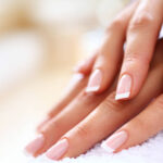 Closeup of nicely manicured female fingernails. One hand is placed on top of other, both on a white towel. Very nice french manicure with transparent nail paint. Blurry beige background. Copy space.